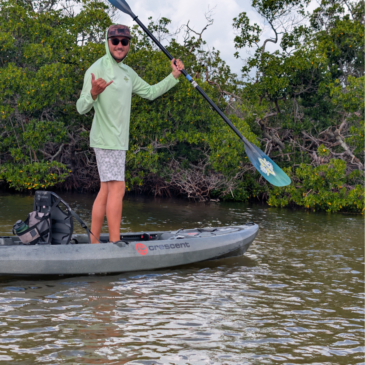 Person in light green hoodie standing on a kayak with a paddle near green shrubs.